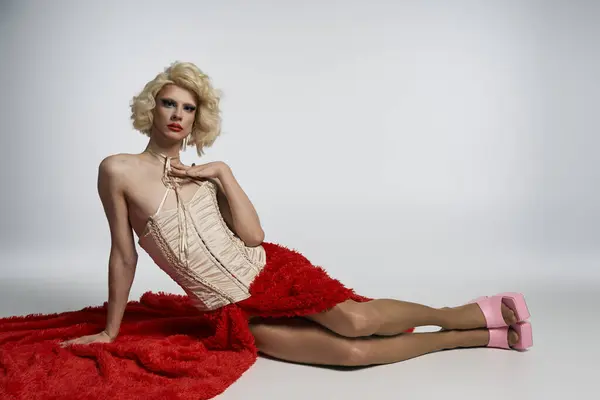 Blonde drag queen in red skirt and corset, legs stretched out, against grey backdrop — Stock Photo