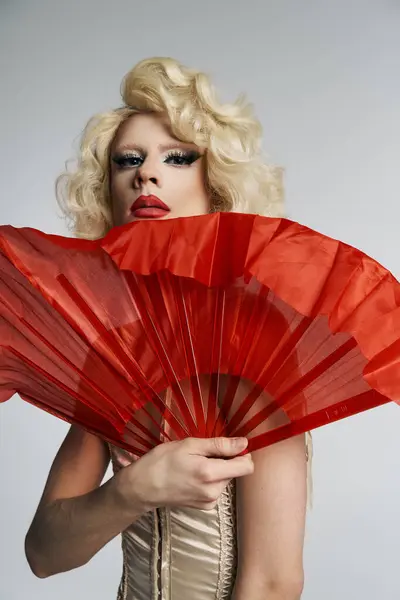 A drag queen in a blonde wig holds a red fan in front of her face, posing against a grey backdrop. — Stock Photo