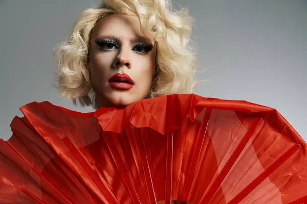 Una drag queen con trucco audace e una parrucca bionda posa con un grande ventilatore rosso contro uno sfondo grigio. — Foto stock
