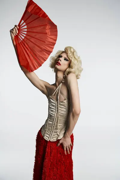 Una drag queen in una parrucca bionda e gonna rossa posa con un grande ventilatore rosso davanti a uno sfondo grigio. — Foto stock