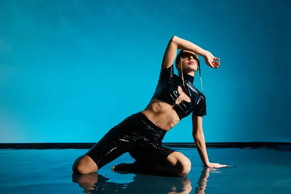 A seductive woman in a black latex outfit poses on a reflective blue surface, her body creating a striking contrast against the backdrop. — Stock Photo