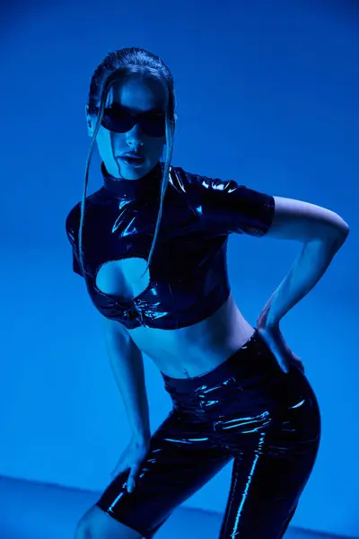 A young woman poses in a black latex crop top and shorts, illuminated by a blue light. — Stock Photo