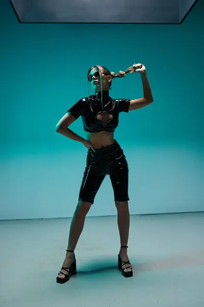 A young woman in a black latex crop top and shorts poses against a teal backdrop, looking confidently to the side. — Stock Photo