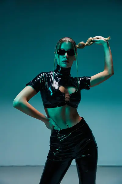 A young woman in black latex pants and a crop top poses with a hand in her hair. The background is a teal green color. — Stock Photo