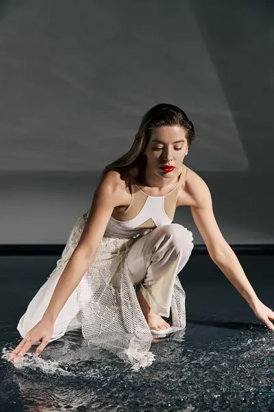 A young woman in a white outfit poses on a grey background, her hand reaching out to the water. — Stock Photo