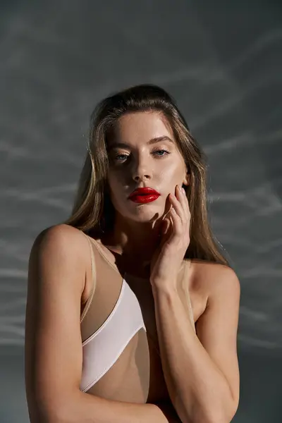 A young woman with long hair, wearing a white outfit, poses with her hand on her cheek against a grey background. — Stock Photo