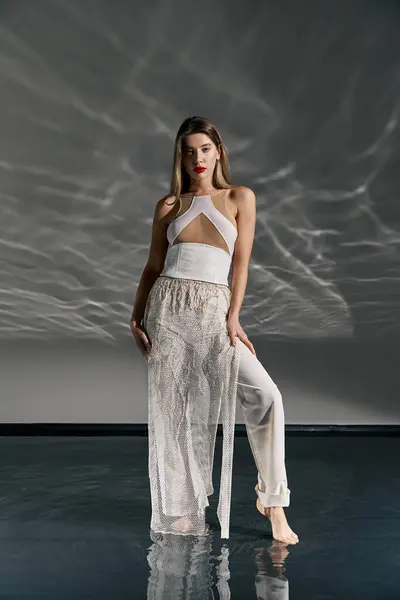 A young woman in a white outfit poses on a grey background, with a reflective surface that makes her appear to be walking on water. — Stock Photo