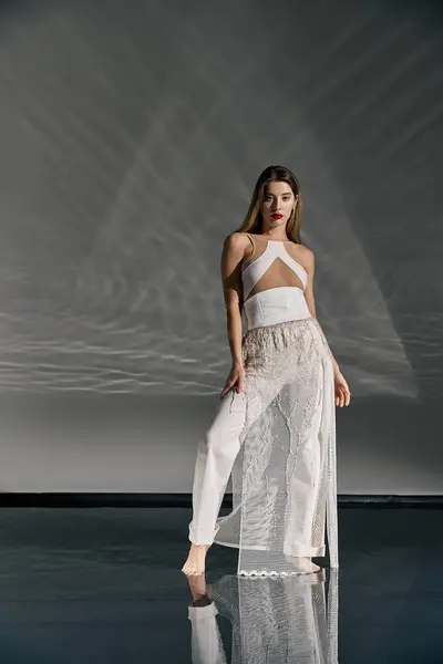 A young woman in a white outfit poses on a reflective floor against a grey backdrop. — Stock Photo