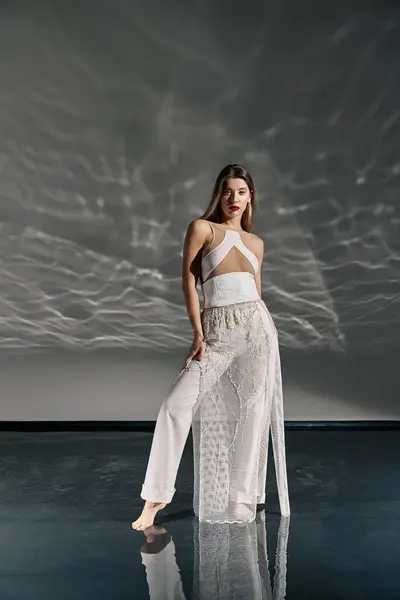 A young woman in a white outfit poses on a grey background with a white, wave-like pattern projected onto the wall. — Stock Photo