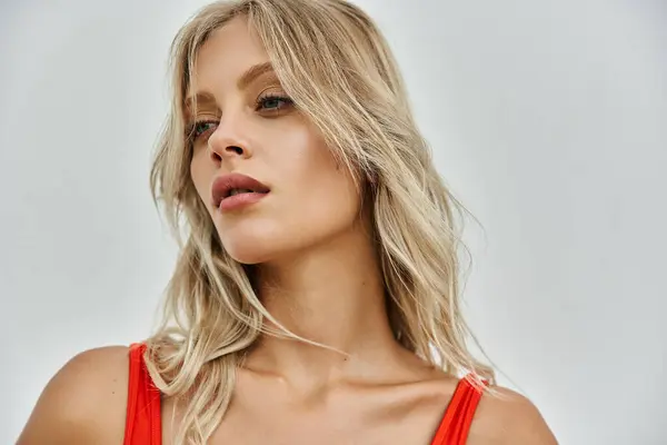A young blonde woman with a bright orange swimsuit poses against a grey background. Her hair is flowing and her expression is confident. — Stock Photo