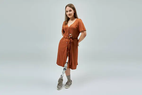 A woman with a prosthetic leg stands confidently in a rust-colored dress against a plain background. — Stock Photo