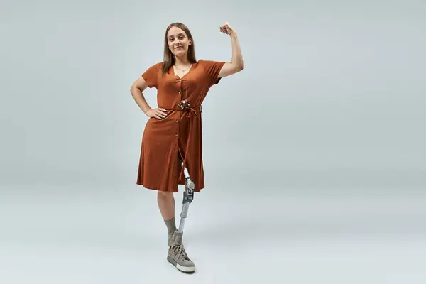 Una mujer con un vestido marrón está de pie con el brazo derecho levantado, mostrando su pierna protésica. — Stock Photo