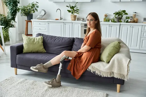 Une femme en robe brune s'assoit sur un canapé avec sa jambe prothétique étendue. — Stock Photo