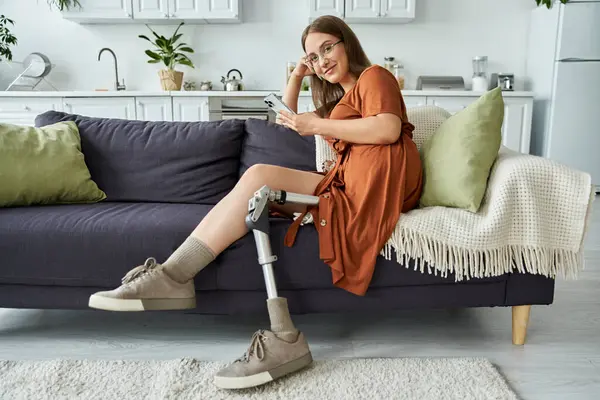 A woman with a prosthetic leg sits on a couch, smiling as she looks at her phone. — Stock Photo