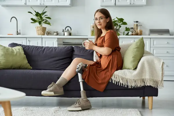 A woman sits on a couch, holding a mug in a relaxed pose, her prosthetic leg visible. — Stock Photo