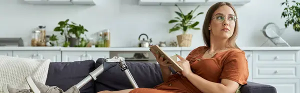 A woman with a prosthetic leg sits on a couch, holding a book and looking thoughtful. — Stock Photo