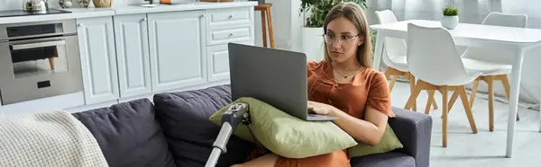 A woman sits on a sofa with a prosthetic leg, using a laptop while looking intently at the screen. — Stock Photo
