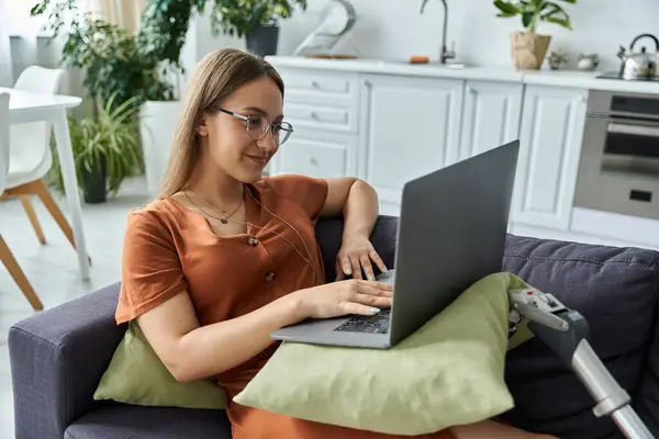 Uma mulher com uma perna protética senta-se em um sofá, sorrindo enquanto trabalha em um laptop. — Fotografia de Stock