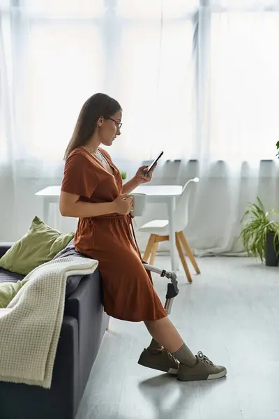 A woman with a prosthetic leg sits on a sofa, looking at her phone and holding a mug. — Stock Photo