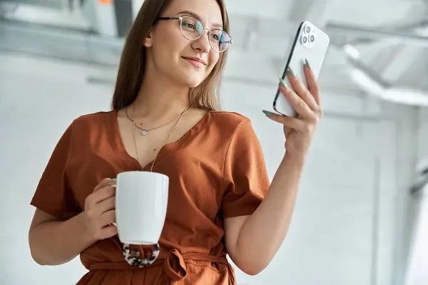 Eine Frau lächelt, während sie einen Kaffeebecher in der Hand hält und auf ihr Handy blickt. — Stockfoto