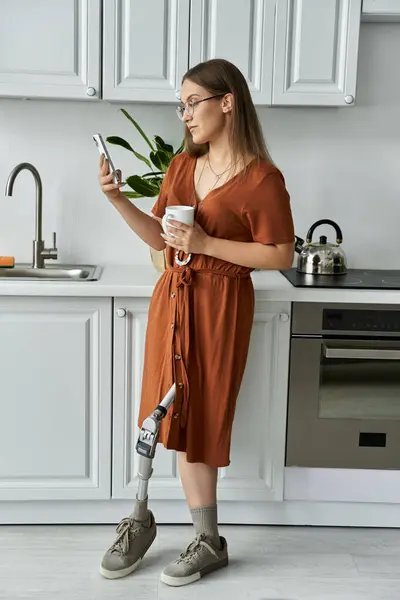 A woman in an orange dress stands in a kitchen, looking at her phone while holding a cup of coffee. — Stock Photo