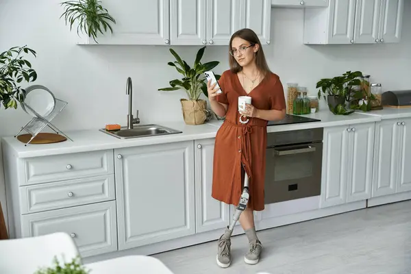 Eine Frau mit Beinprothese steht in einer modernen Küche und hält ein Telefon und eine Tasse Kaffee in der Hand. — Stockfoto