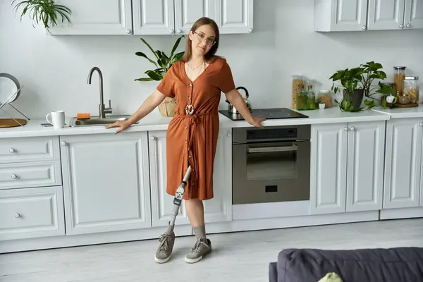 Une femme en robe brune sourit en toute confiance dans sa cuisine, sa jambe prothétique visible. — Photo de stock