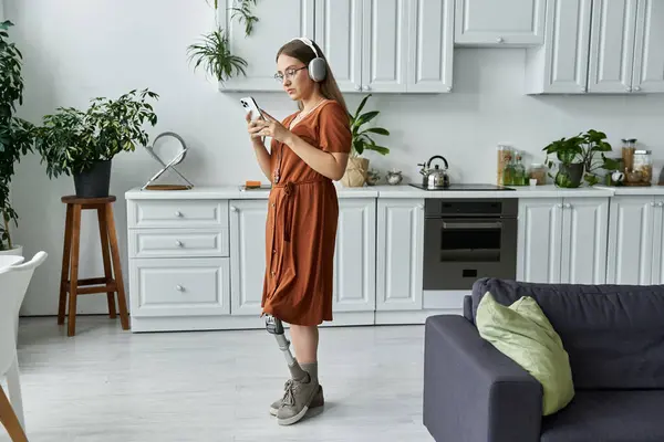 A woman with a prosthetic leg uses her phone while wearing headphones in a kitchen. — Stock Photo