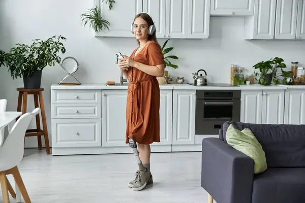 A woman with a prosthetic leg stands in a modern kitchen, listening to music. — Stock Photo