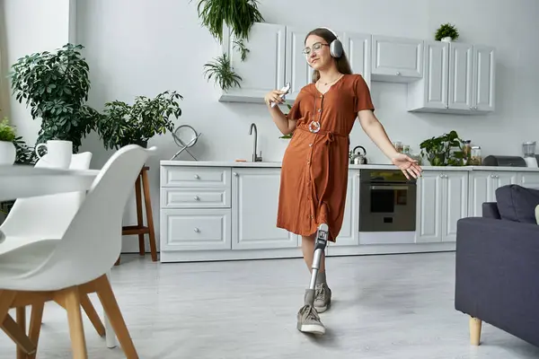 A woman with a prosthetic leg dances in a kitchen, wearing a dress and headphones. — Stock Photo