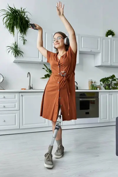 Uma mulher em um vestido marrom dança em sua cozinha enquanto usa fones de ouvido e segurando um telefone. — Fotografia de Stock