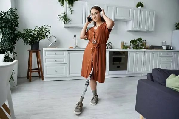 A woman in a brown dress and headphones dances with a prosthetic leg in a bright kitchen. — Stock Photo