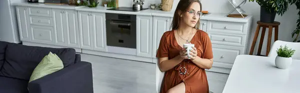 Eine Frau sitzt an einem Tisch, hält einen Becher in der Hand und schaut aus dem Fenster. — Stockfoto