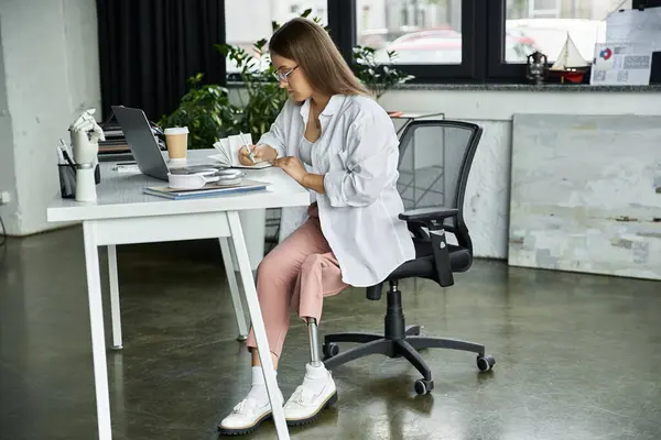 Una donna con una gamba protesica si siede a una scrivania, lavorando a un progetto. — Foto stock