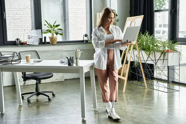 A woman with a prosthetic leg leans against a desk while working on a laptop. — Stock Photo