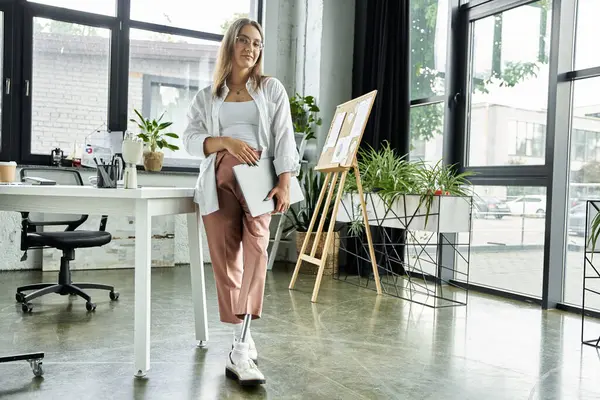 Eine Frau mit Beinprothese lehnt an einem Schreibtisch in einem Büroraum und hält einen Laptop in der Hand. — Stockfoto