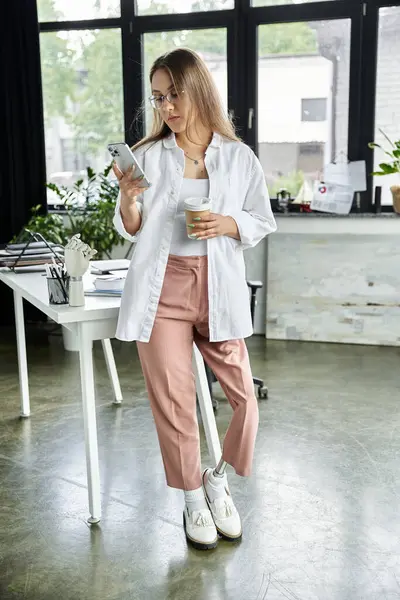 Woman with prosthetic leg scrolls phone, holds coffee cup by desk — Stock Photo