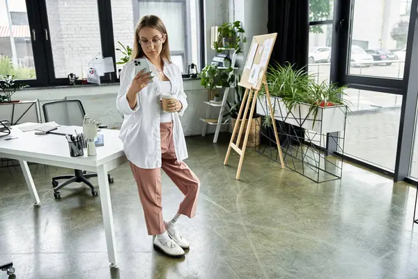Woman in white shirt and pink pants, leaning against desk, checks phone and holds coffee cup — Stock Photo