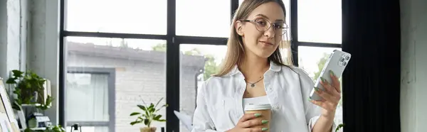 A woman in glasses stands near a window, looking at her phone while holding a cup of coffee. — Stock Photo