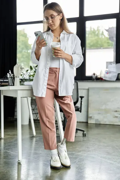 A woman with a prosthetic leg stands by a desk in an office, checking her phone. — Stock Photo