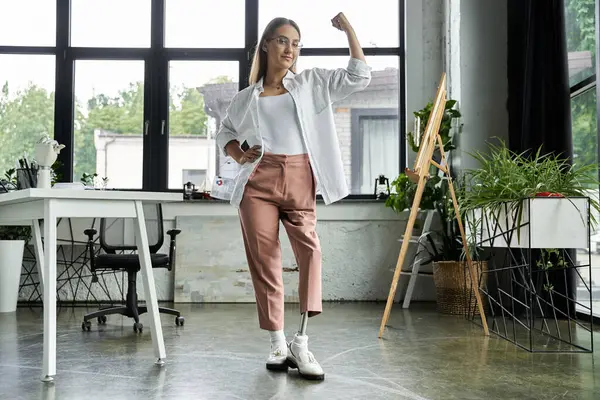 Mujer con la pierna protésica se para con confianza en la oficina, brazo levantado en fuerza - foto de stock