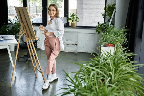 A young woman with a prosthetic leg stands by an easel in a studio, surrounded by plants. — Stock Photo