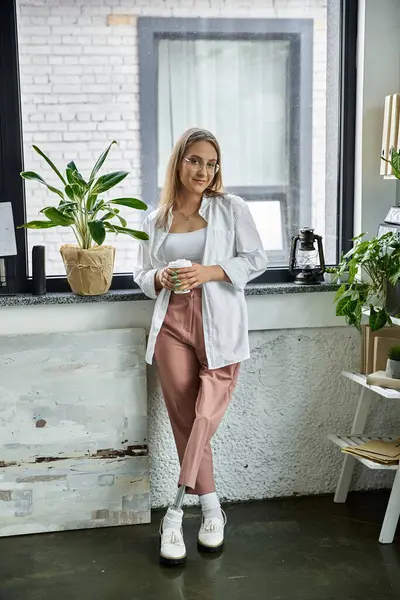 A woman with a prosthetic leg stands by a window, holding a cup and looking out. — Stock Photo