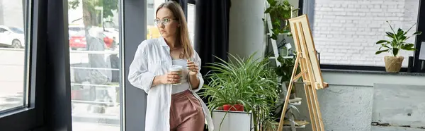 Woman near window, holding coffee, with easel and plants — Stock Photo