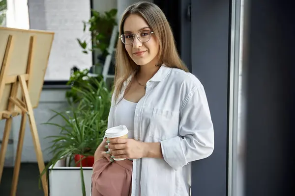 A woman in a white shirt holds a coffee cup and looks directly at the camera, showcasing confidence. — Stock Photo