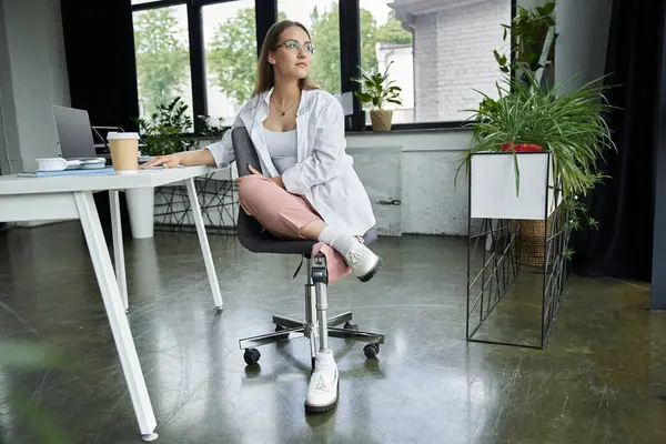 A woman with a prosthetic leg sits at a desk, gazing out the window. — Stock Photo
