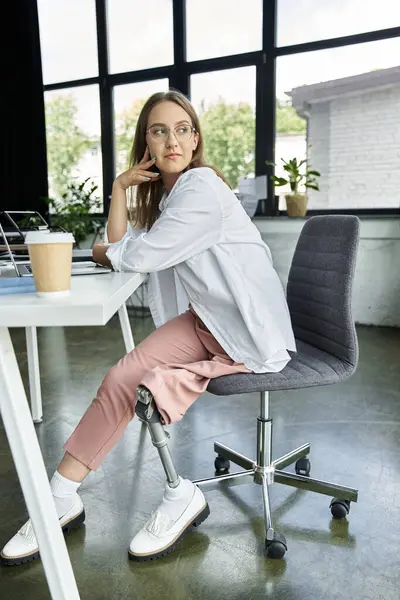 A woman with a prosthetic leg sits in an office chair, looking out a window. — Stock Photo