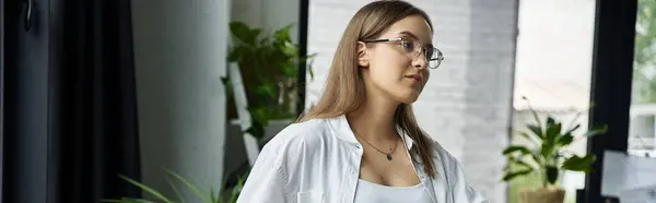 Una mujer con el pelo castaño largo y gafas, vistiendo una camisa blanca, mira por una ventana. — Stock Photo