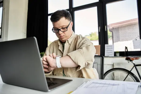 A young man with Down syndrome sits at a desk with a laptop open, checking his watch. — Stock Photo