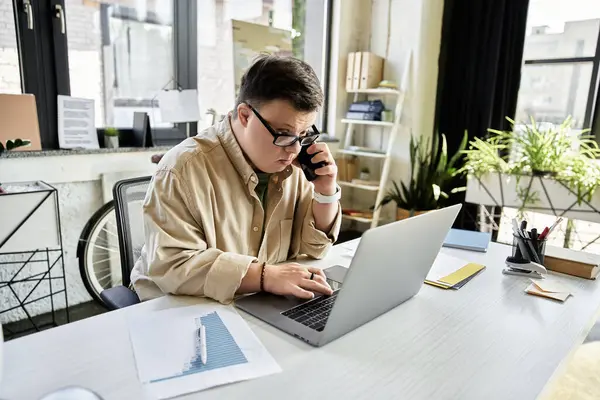 A young man with Down syndrome uses a laptop and phone at his desk. — Stock Photo